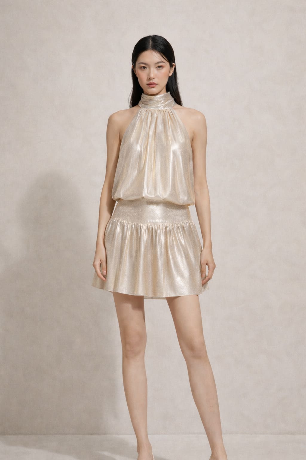 Studio portrait of a woman wearing a champagne halter-neck metallic mini dress with a softly draped bodice and flared skirt against a neutral beige background