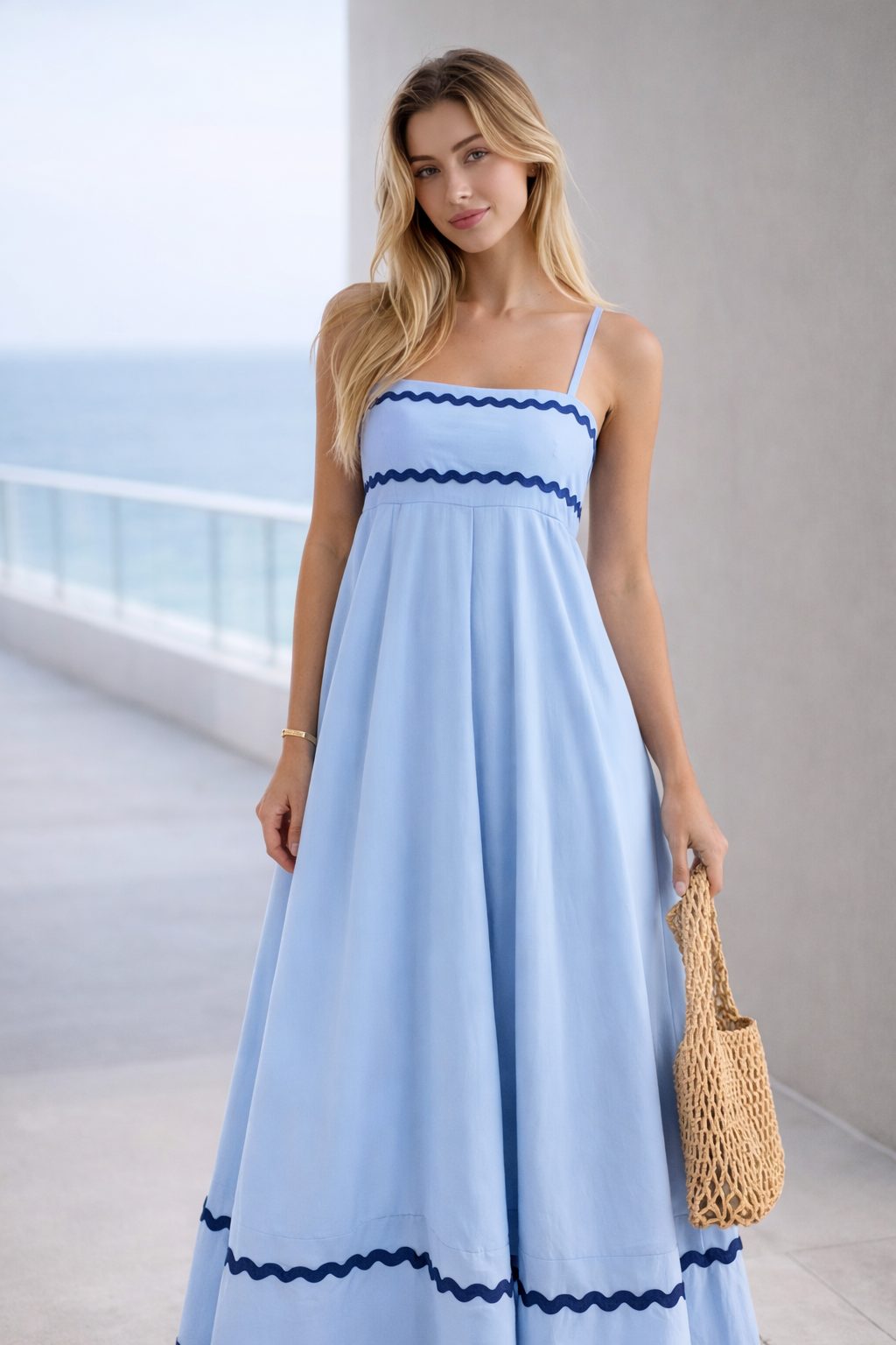 Woman wearing a light blue sleeveless maxi dress with contrasting wave trim and a woven bag in a modern editorial setting